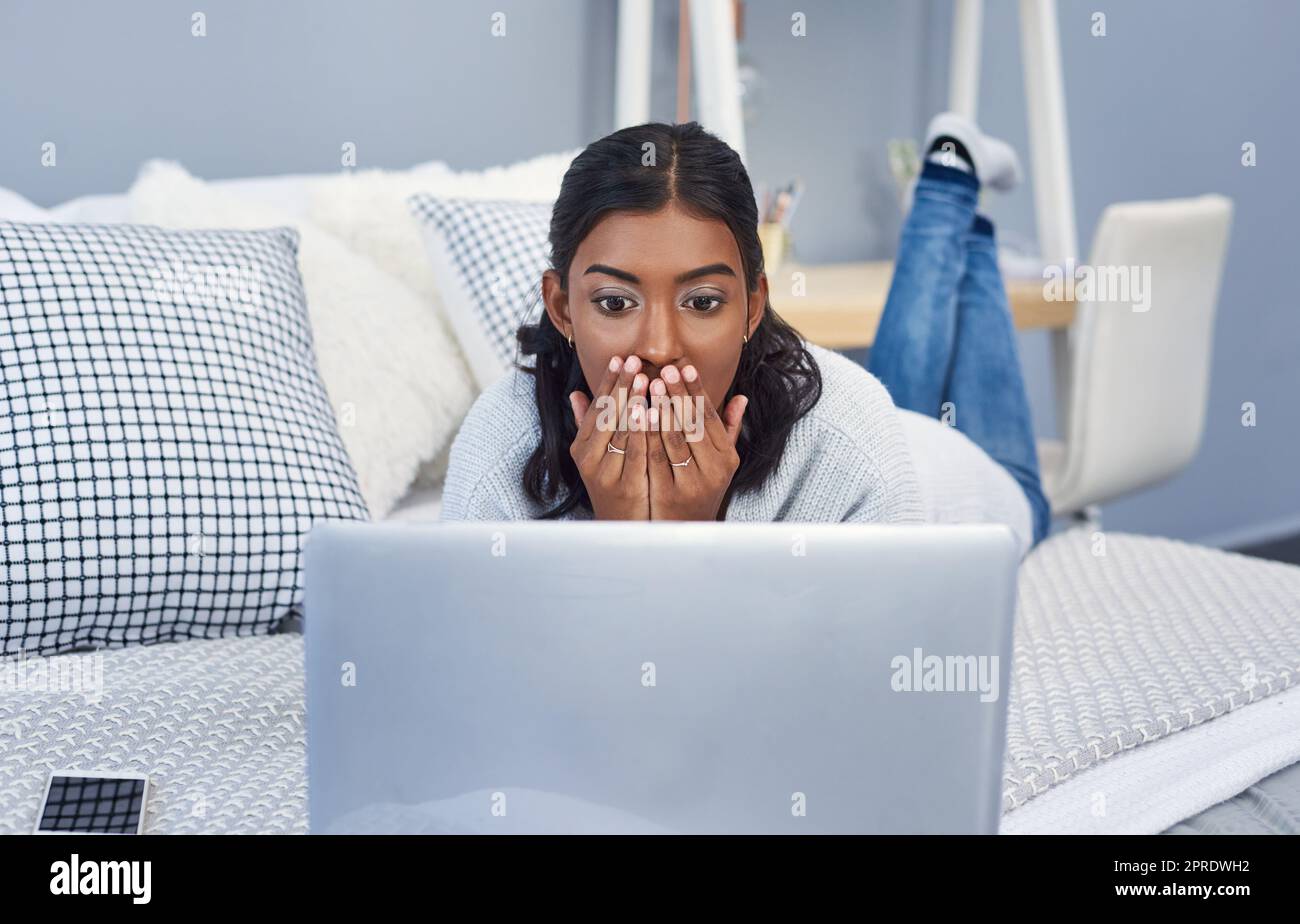 Non riesco a credere a ciò che viene detto online. Una giovane donna attraente che usa un computer portatile mentre si rilassa sul letto nella sua camera da letto a casa. Foto Stock