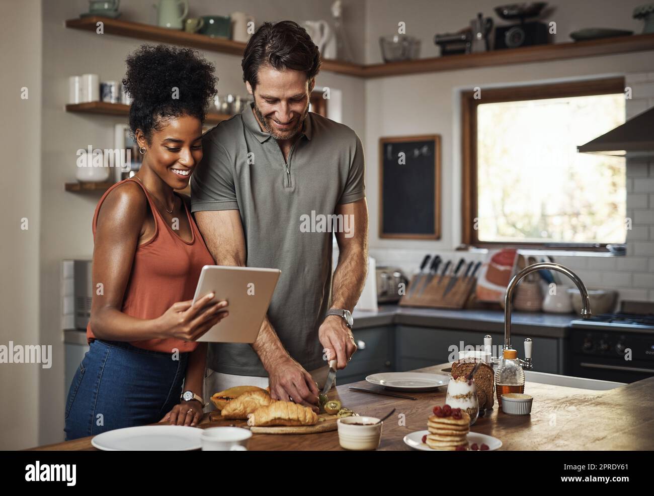 Giovane coppia che cucinano cibo sano insieme seguendo le ricette online su un tablet, passo dopo passo. Marito e moglie felici, allegri e sorridenti che preparano la cena in cucina a casa. Foto Stock