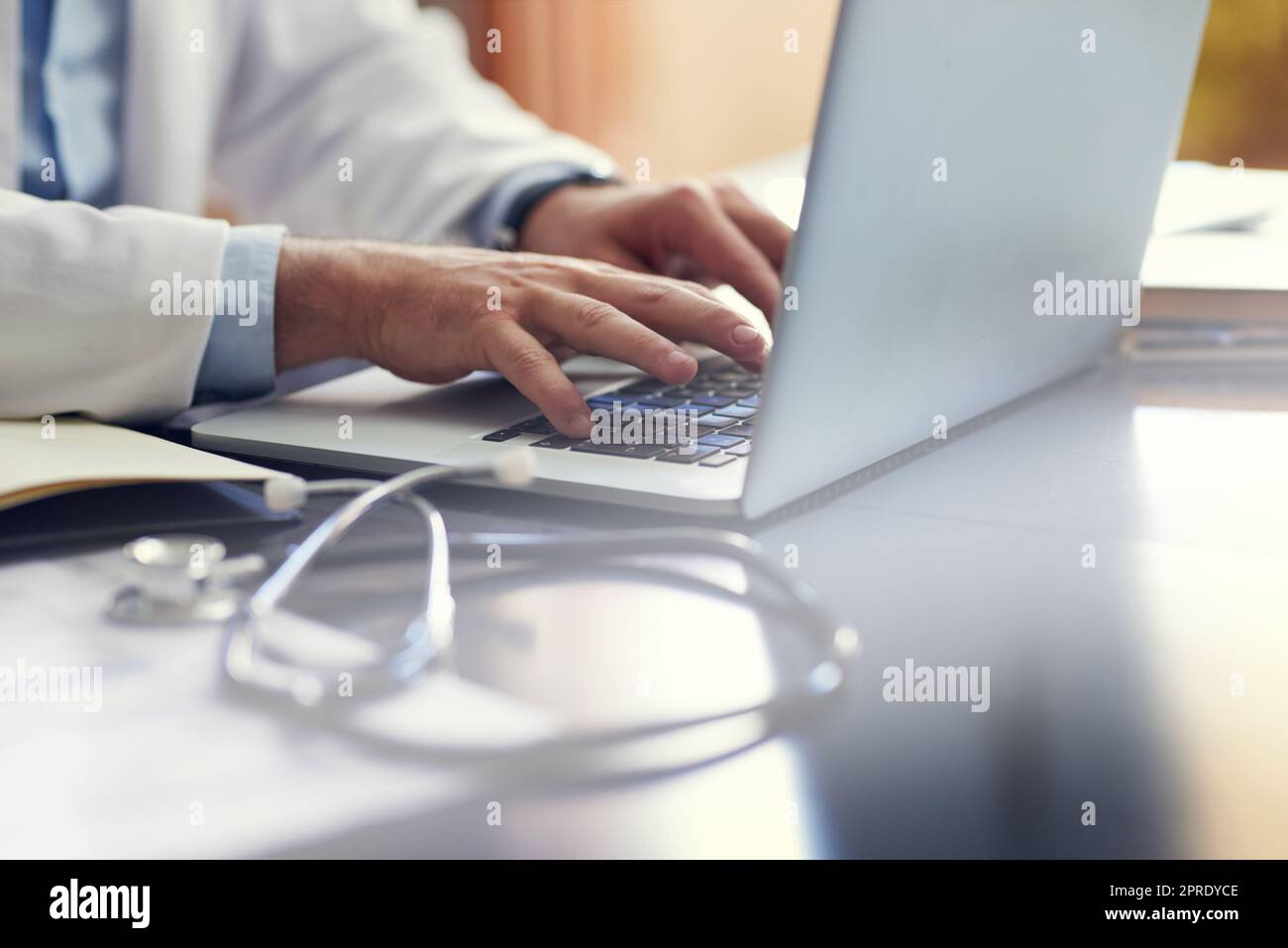 Scrivere le sue relazioni mediche. Un medico maschio irriconoscibile che lavora sul suo computer portatile mentre è seduto in ufficio. Foto Stock