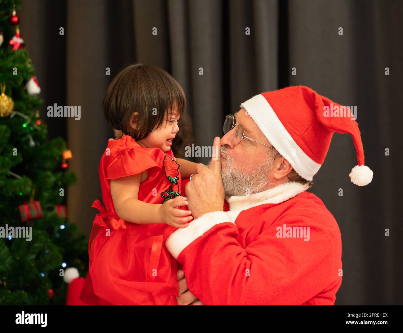 Babbo Natale che indossa il costume rosso che tiene l'indice dalla sua bocca mentre cerca di conforto urlando bambina piangente vicino all'albero di Natale nel roo vivente Foto Stock