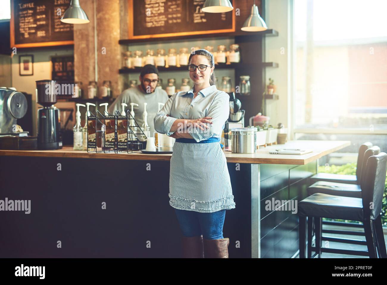 Siamo in grado di soddisfare tutti gli amanti del caffè. Ritratto di una giovane donna che lavora in un bar con la sua collega sullo sfondo. Foto Stock