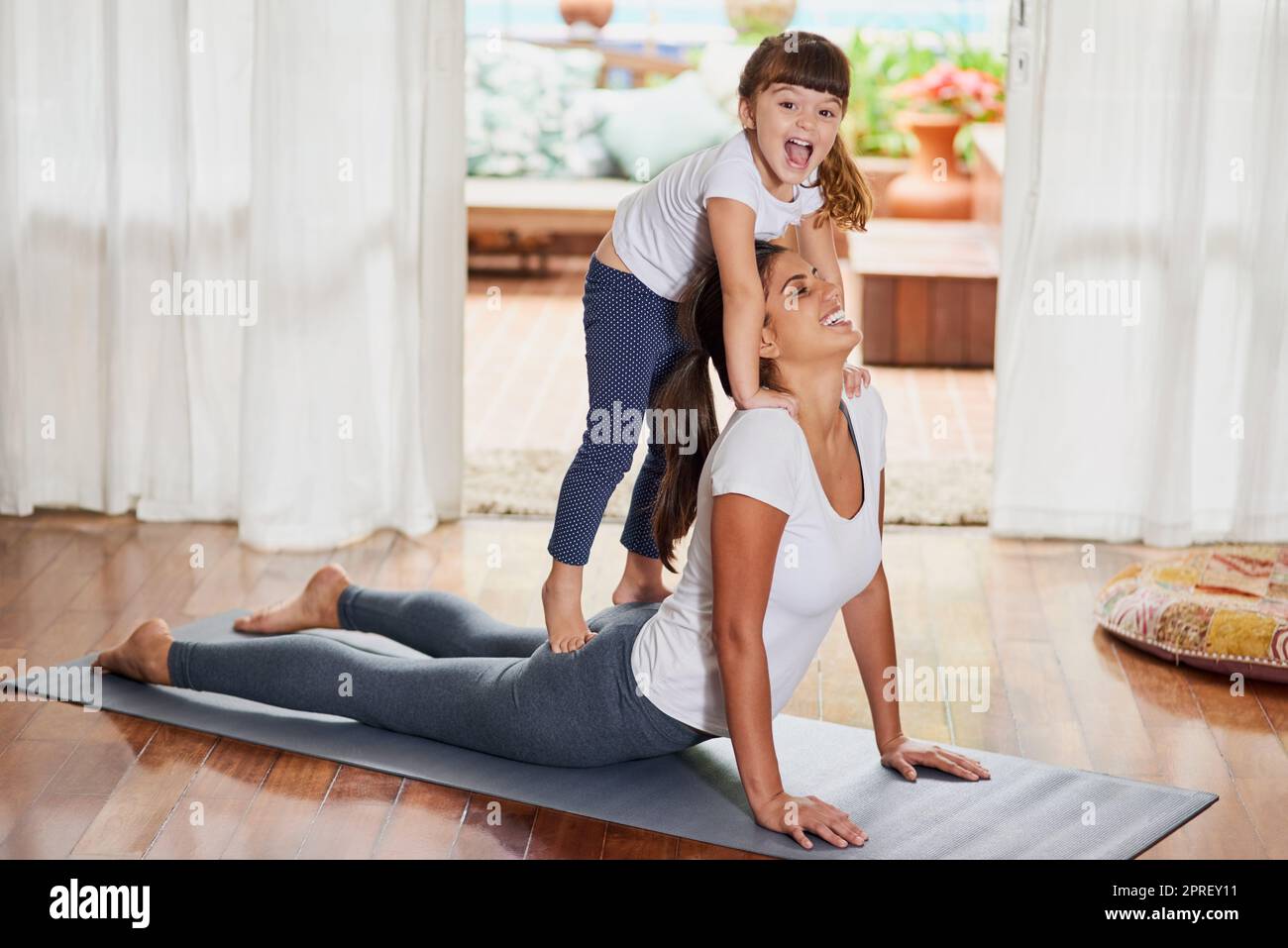 Che posa interessante. Una giovane donna allegra che fa una posa yoga mentre la sua figlia si alza dolcemente sulla schiena a casa. Foto Stock