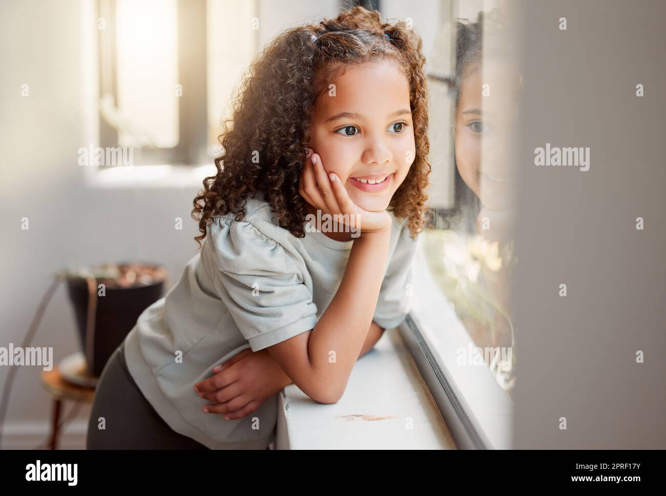 Bambina che sogna di giocare fuori al sole dopo il lockdown, sorridendo e guardando fuori da una finestra. Piccola, carina e felice bambina che aspetta, pensa e sogna ad occhi aperti con la mano sul mento Foto Stock