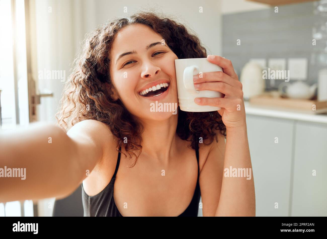 Buon caffè mattutino e selfie sorridendo e tenendo la tazza contro il viso per riscaldarsi mentre si è seduti a casa. Ritratto di una giovane donna allegra che si gode il tempo libero e una bevanda calda Foto Stock