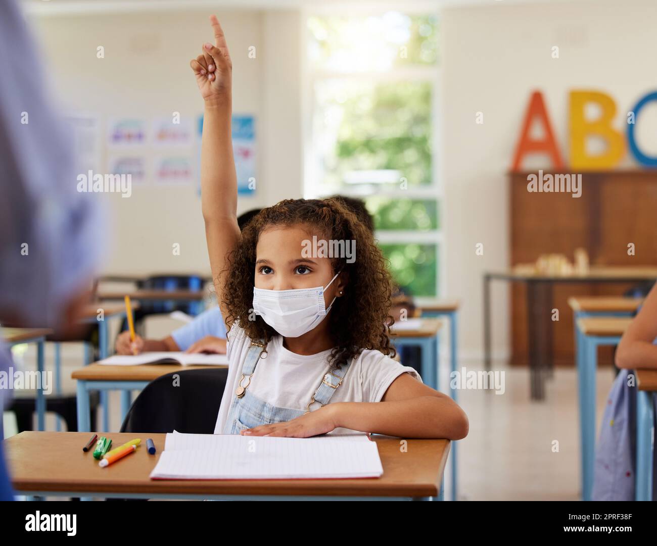 COVID, apprendimento e istruzione con una giovane studentessa che indossa una maschera e alza la mano per fare o rispondere a una domanda in classe a scuola. Bambina che chiede o risponde all'insegnante in classe Foto Stock