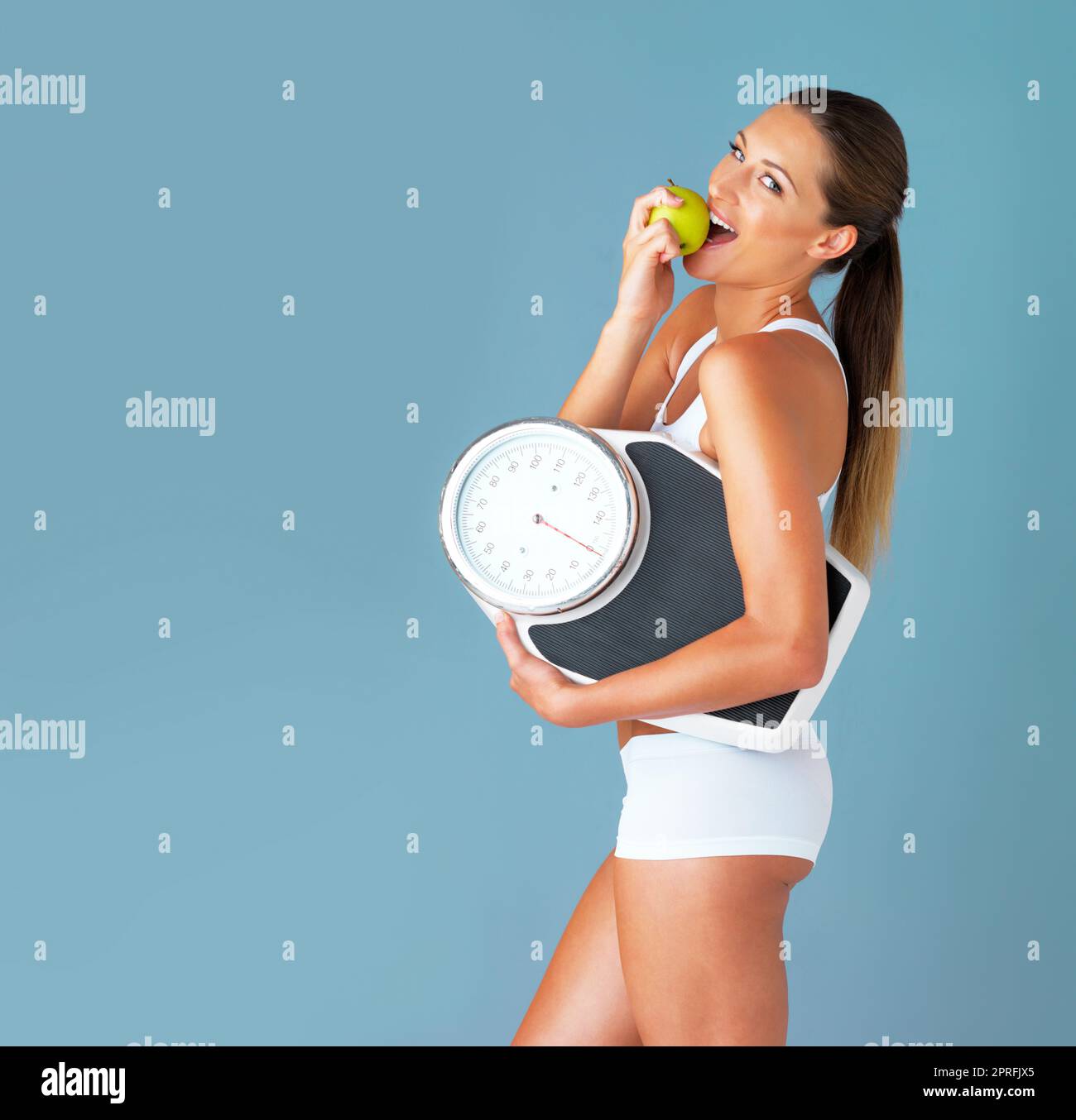 Non è il momento migliore per iniziare a vivere in buona salute. Studio shot di una giovane donna in forma che mangia una mela e tiene una scala su uno sfondo blu. Foto Stock