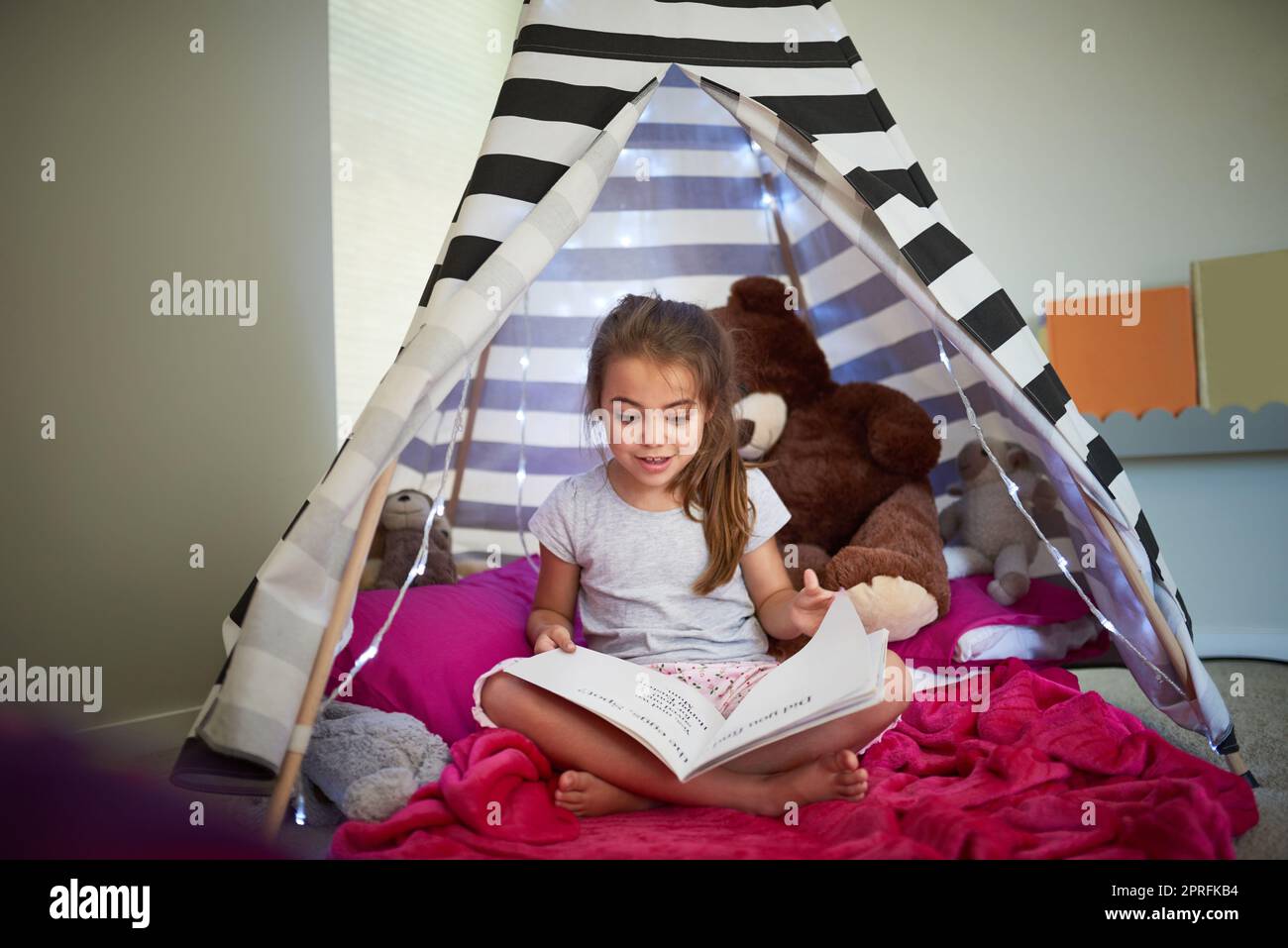 I libri possono contribuire ad alimentare la curiosità e l'immaginazione dei bambini. Una bambina che legge un libro con il suo orsacchiotto in una tenda a casa. Foto Stock