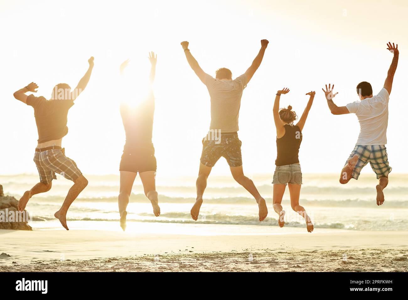 Raggiungi il cielo. Ripresa da dietro di un gruppo di amici non identificabili che saltano insieme sulla spiaggia al tramonto. Foto Stock