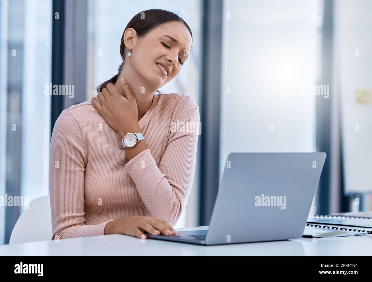 Donna stressante che soffre di dolori al collo che lavora su un notebook in un ufficio moderno. Professionista aziendale con una cattiva postura e un infortunio. Lavoratore SEO aziendale con disagio dovuto alle lunghe ore alla scrivania Foto Stock