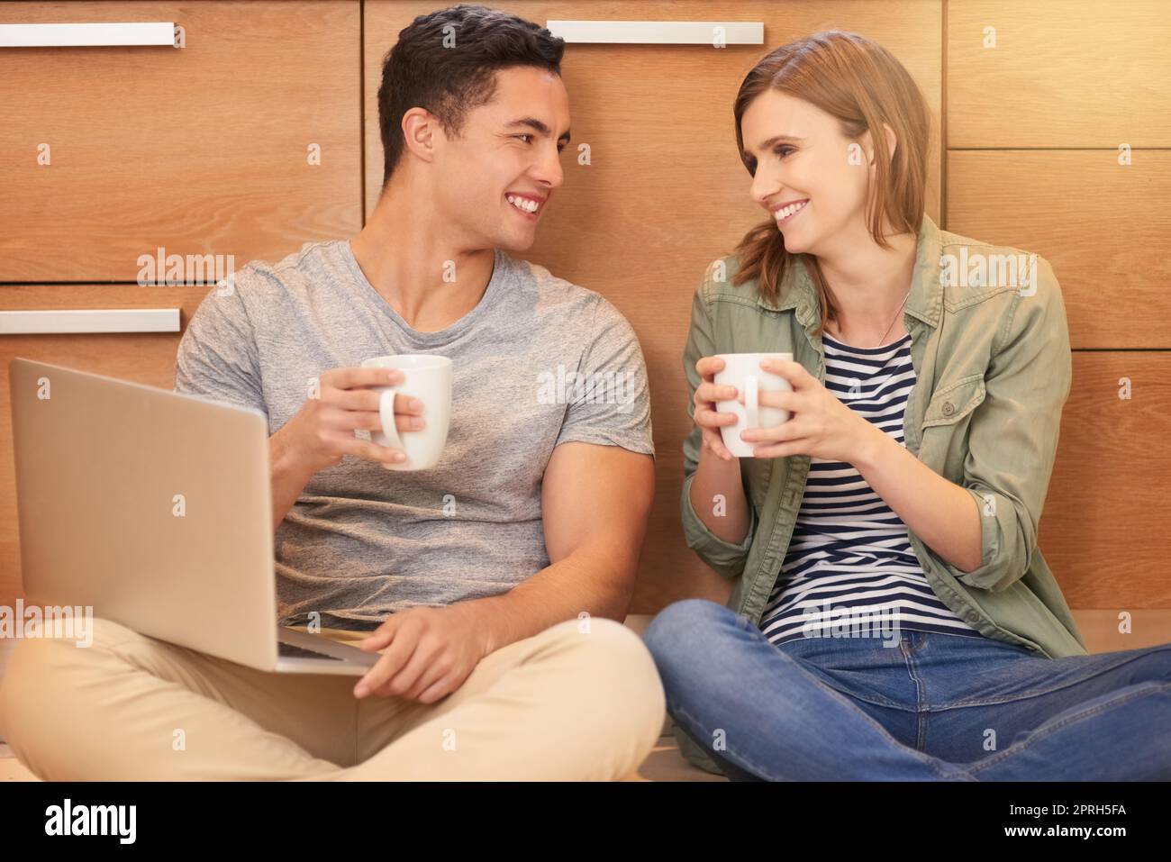È il momento di una pausa caffè. Una giovane coppia felice beve caffè e usa un computer portatile mentre si siede sul pavimento della cucina. Foto Stock