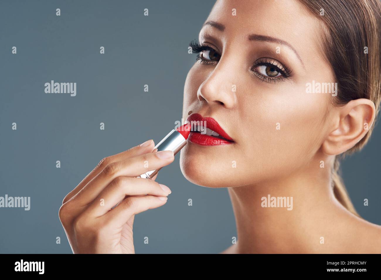 Aggiungere un tocco di glam con rossetto rosso. Studio ritratto di una bella giovane donna mettere su rossetto rosso su uno sfondo grigio. Foto Stock