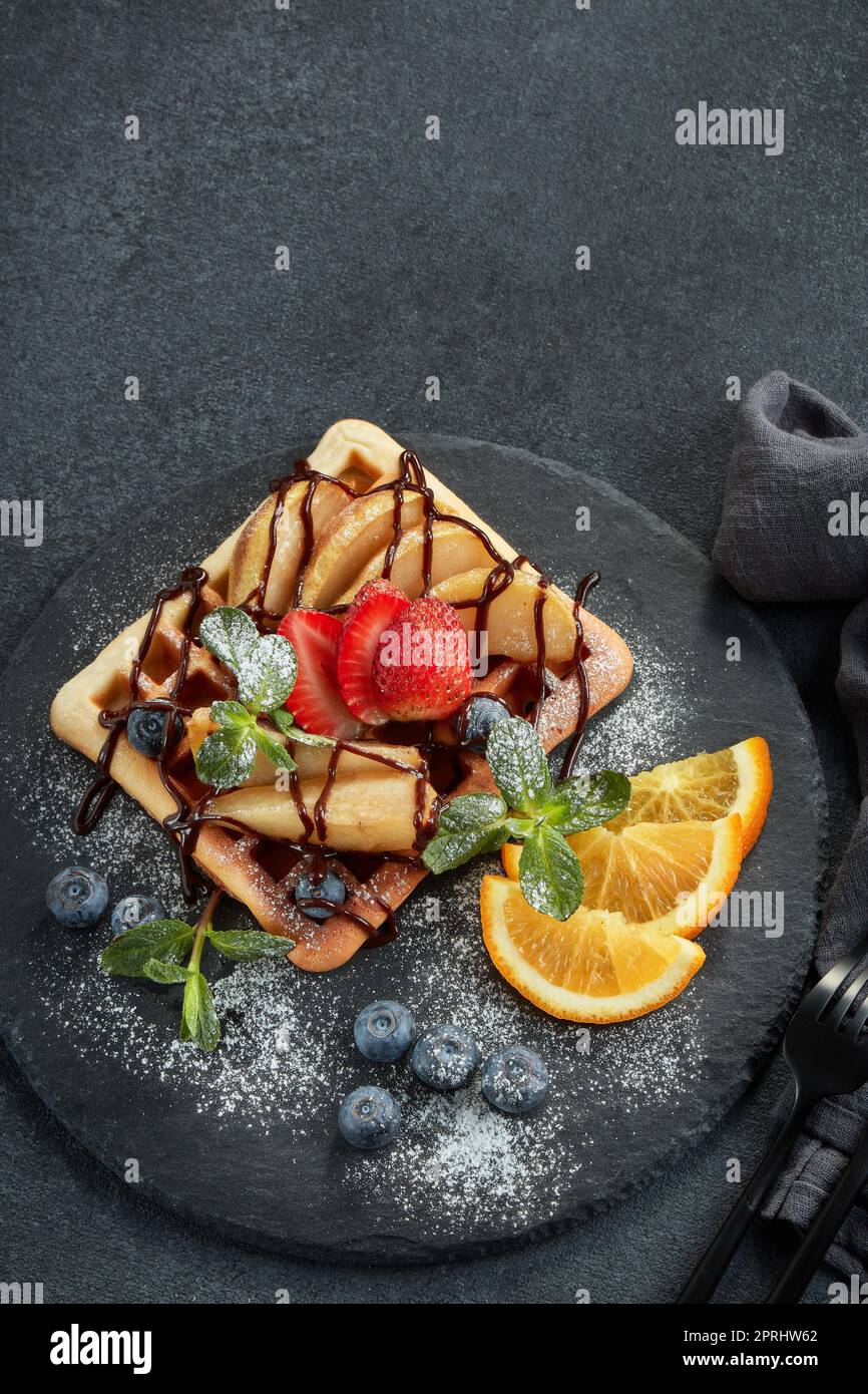 Deliziosi waffle con pera caramellata, frutti di bosco, menta, zucchero a velo e salsa dolce sul piano di servizio nero su sfondo grigio. Dessert. Serving foo Foto Stock