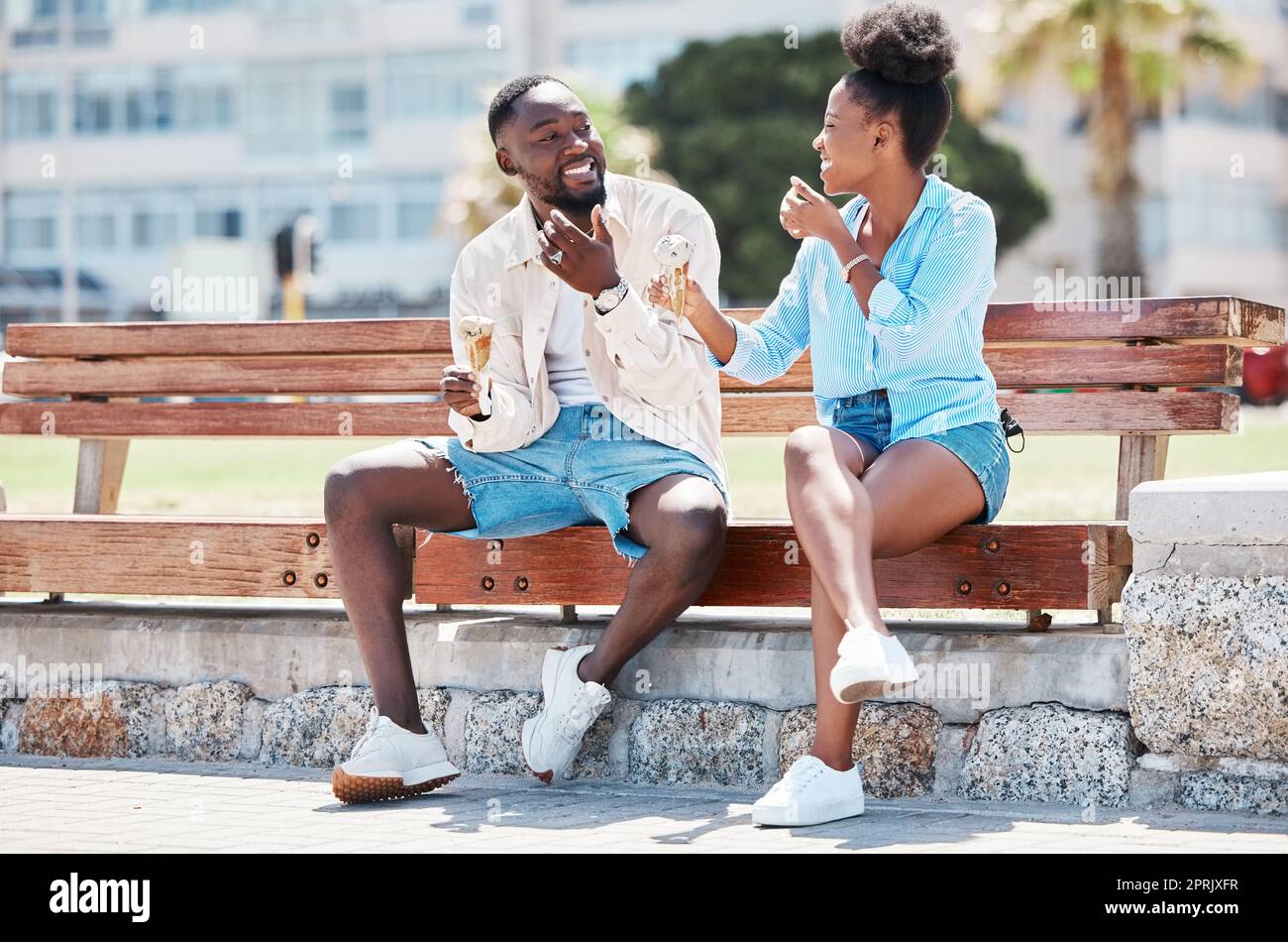 Felice coppia nera che mangia un gelato su una panchina da spiaggia insieme, sorridendo mentre si legano e ridono. Giovane uomo e donna afro-americano che si gode il loro romanticismo estivo, il tempo libero e la loro relazione Foto Stock