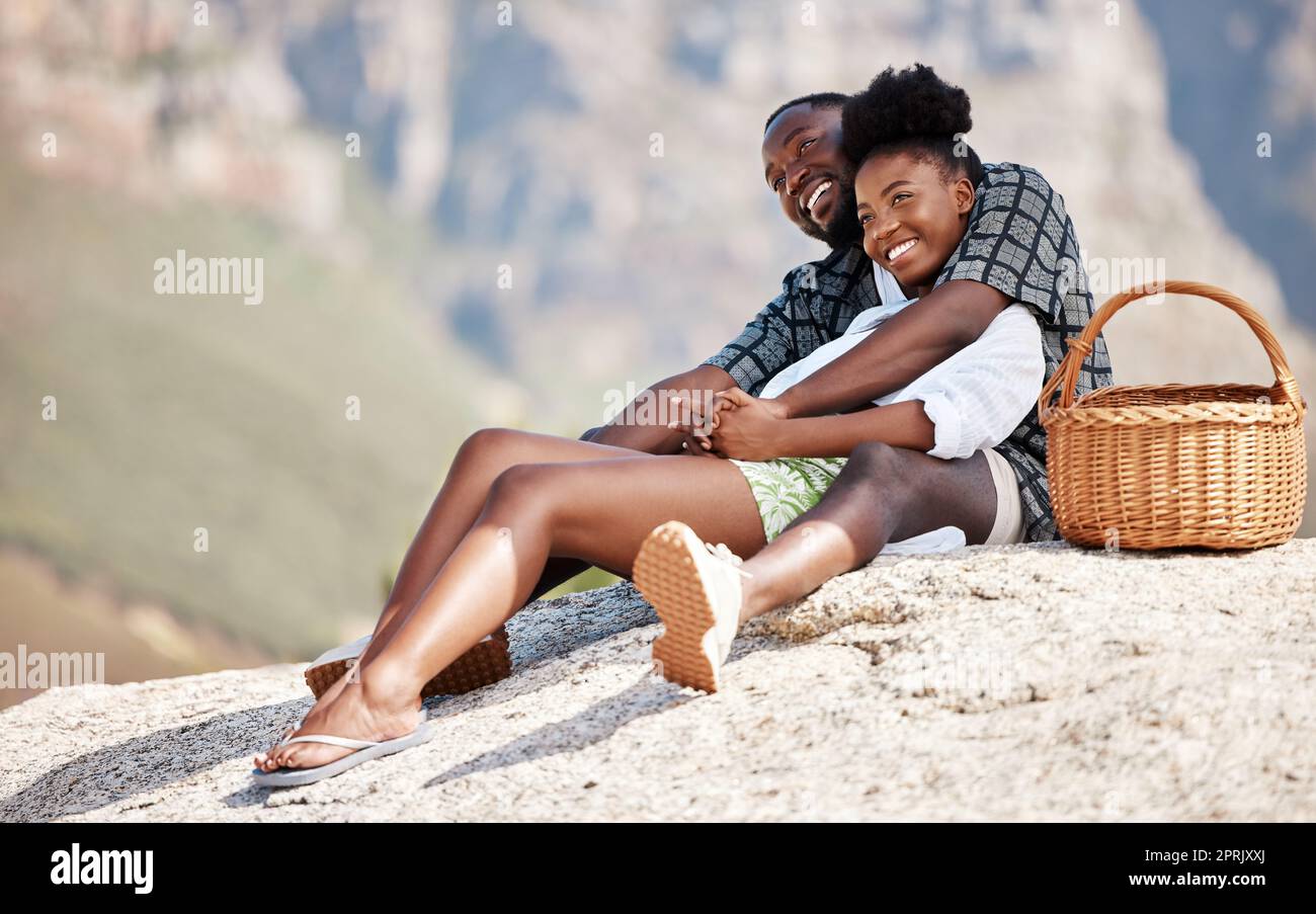 Coppia nera, picnic e amore con un sorriso su una roccia con sfondo montano offuscato in Sudafrica. Rilassatevi, uomini e donne nella natura, vacanze o vacanze estive, fiducia e sicurezza insieme. Foto Stock