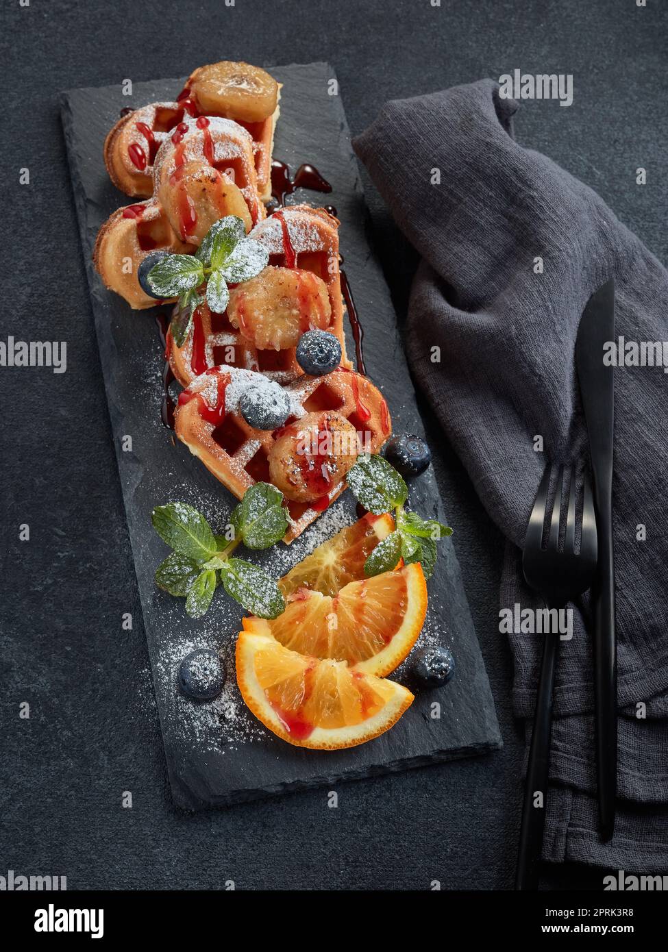 Deliziosi waffle con pera caramellata, frutti di bosco, menta, arancia e salsa dolce sul piano di servizio nero su sfondo grigio. Vista dall'alto. Pasto dolce. Dessert Foto Stock