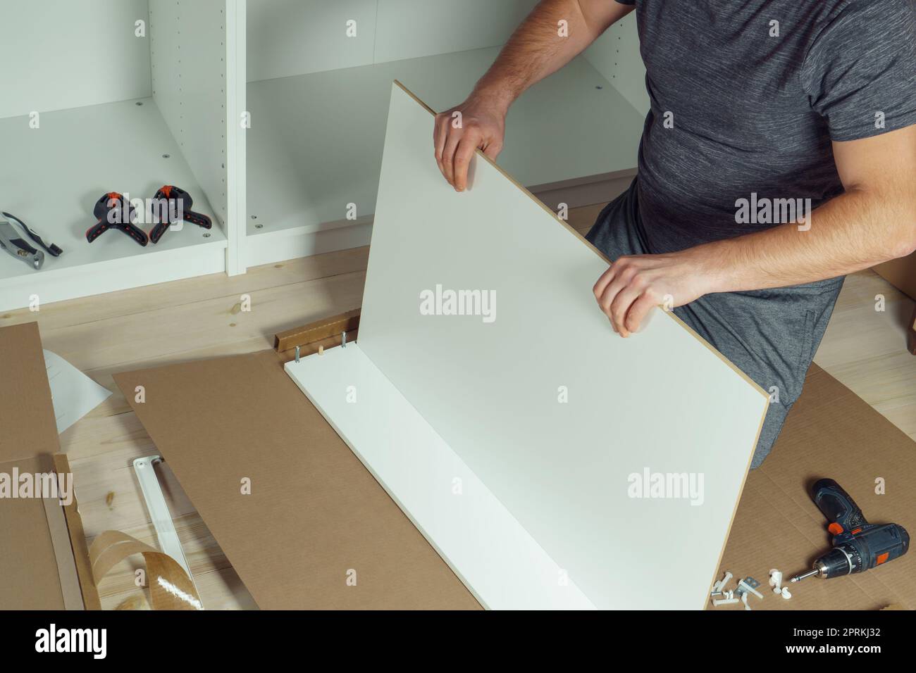 Le mani maschili tengono e inseriscono il fondo del cassetto bianco, montando la comò o il guardaroba sul pavimento. Il cacciavite elettrico e i morsetti sono affiancati. Auto-assemblaggio Foto Stock