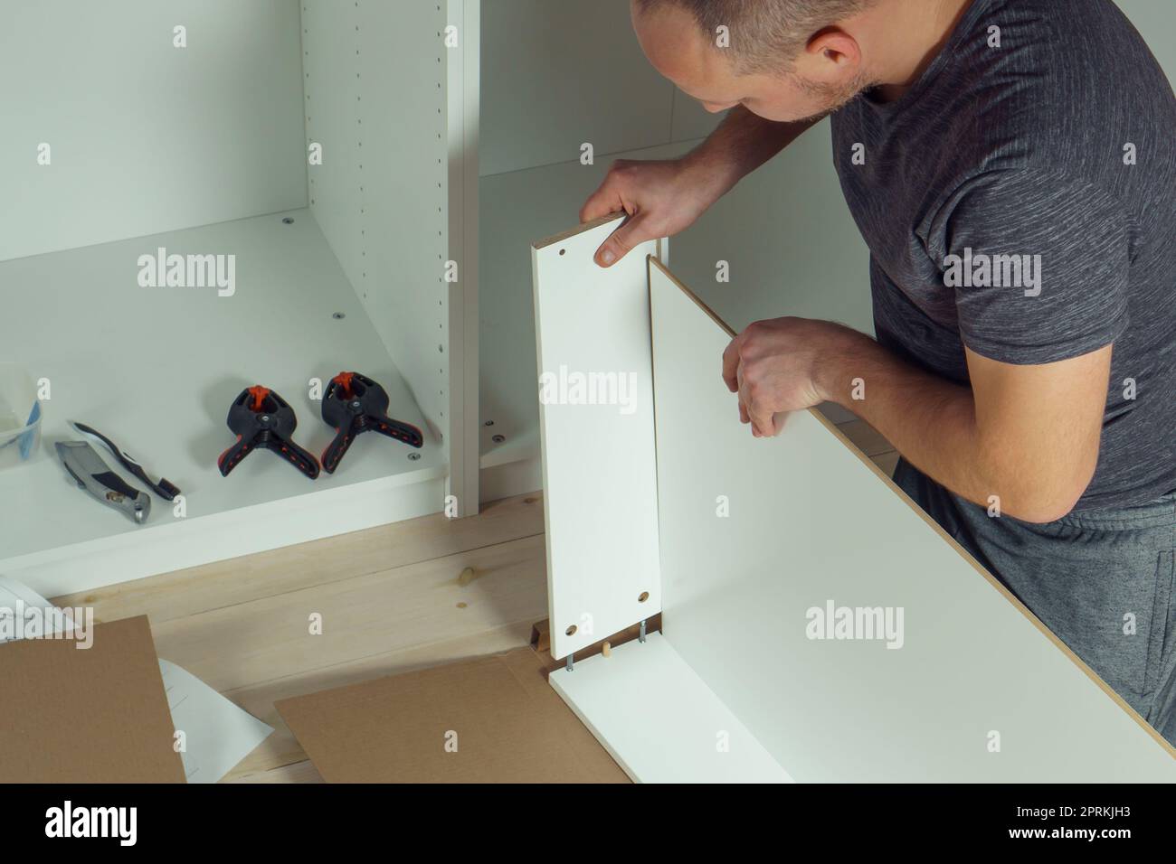 Le mani maschili tengono e inseriscono il fondo del cassetto bianco, montando la comò o il guardaroba sul pavimento. Il cacciavite elettrico e i morsetti sono affiancati. Auto-assemblaggio Foto Stock