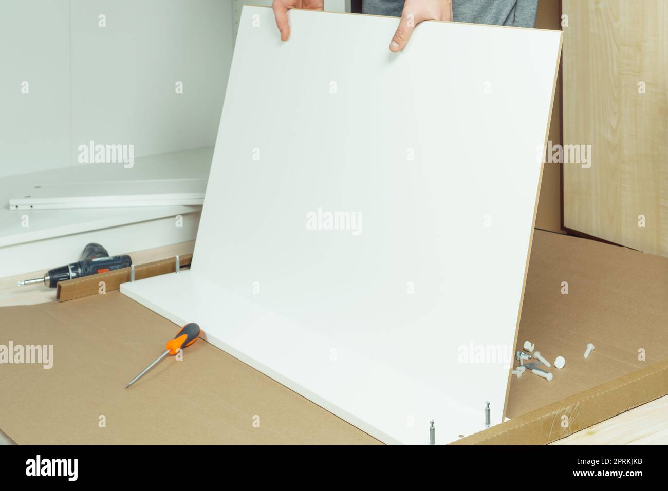 Le mani maschili tengono e inseriscono il fondo del cassetto bianco, montando la comò o il guardaroba sul pavimento. Il cacciavite elettrico e le viti sono affiancati. Auto-assemblaggio Foto Stock
