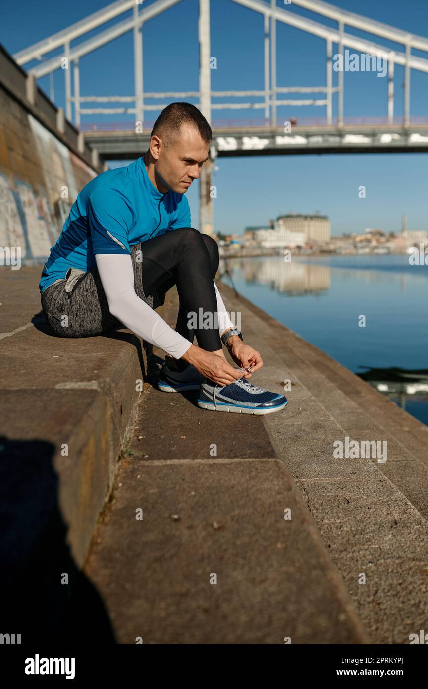 Atleta runner uomo legando lacci su scarpe seduti sulle scale prima di allenarsi sulla riva del fiume della città. Allenamento mattutino per la perdita di peso e la costruzione del corpo Foto Stock