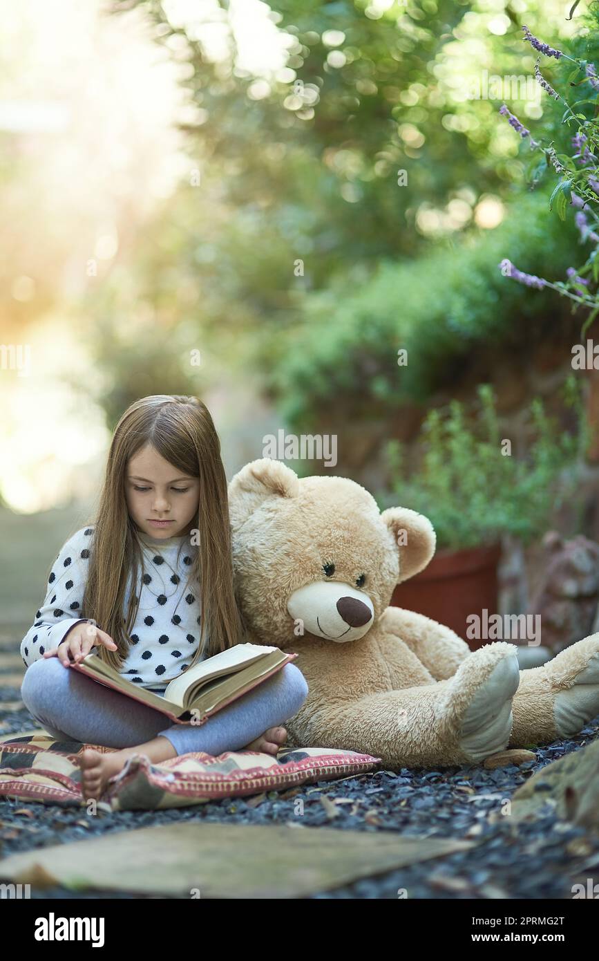 Perso nella meraviglia della sua storia preferita. Una bambina che legge un libro con il suo orsacchiotto accanto a lei. Foto Stock