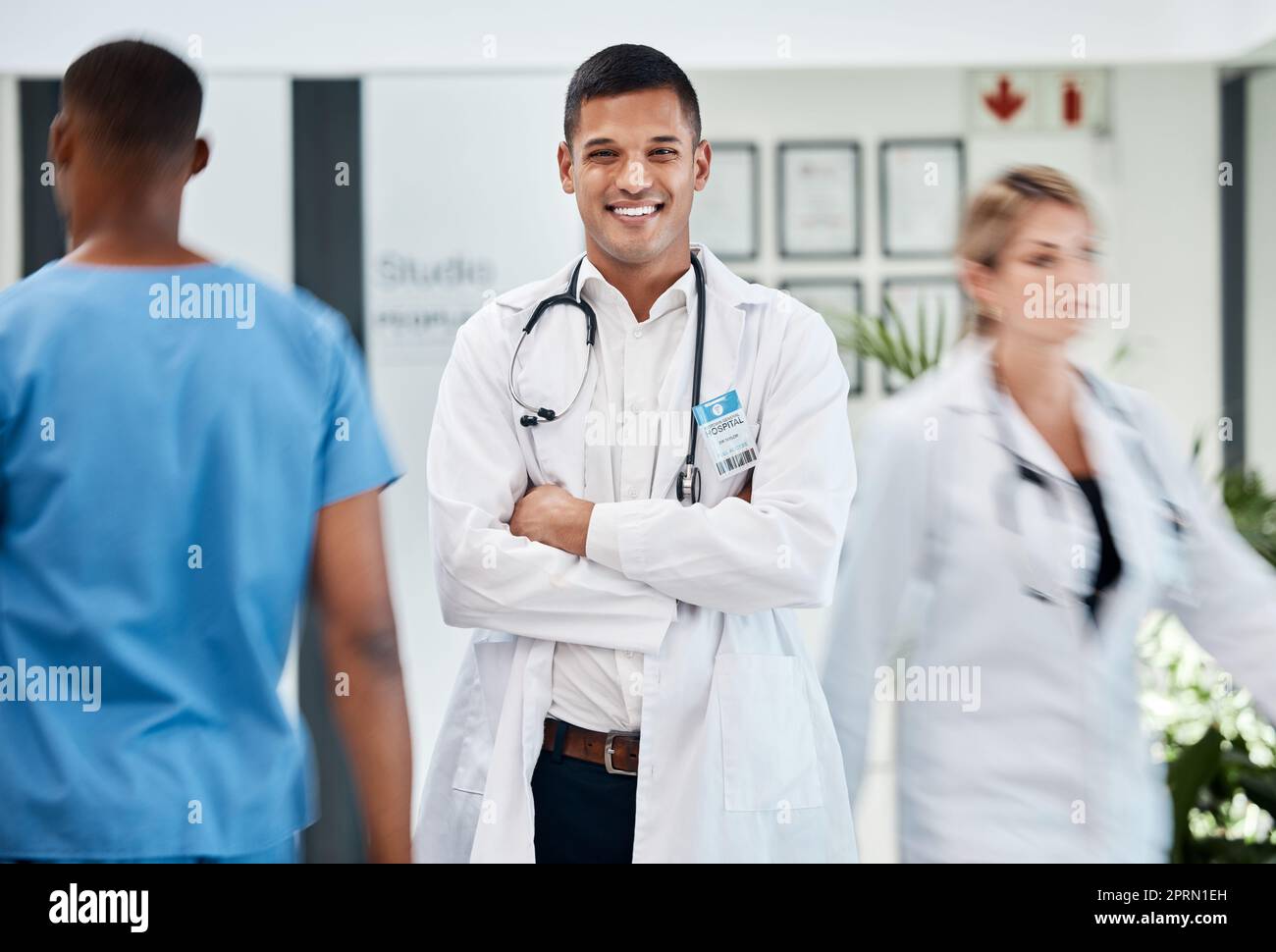 Felice, medico e medico in ospedale con sorriso e sicurezza, braccia incrociate in corridoio affollato. Maschio, chirurgo e medico specialista sanitario in ospedale. Personale, tirocinio clinico. Foto Stock