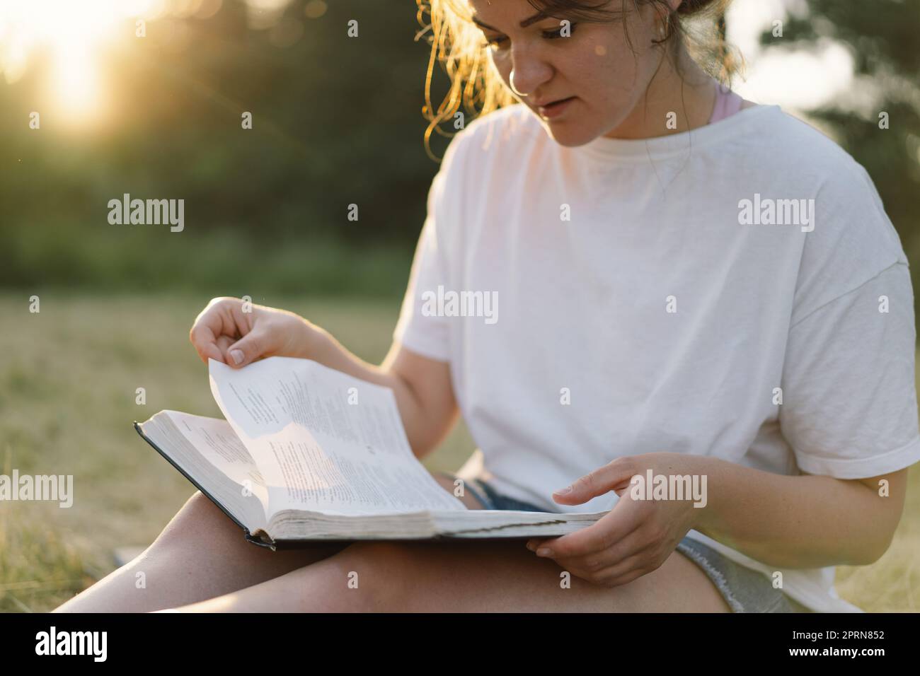 La donna cristiana tiene la bibbia nelle sue mani. Leggere la Sacra Bibbia in un campo durante il bellissimo tramonto. Foto Stock