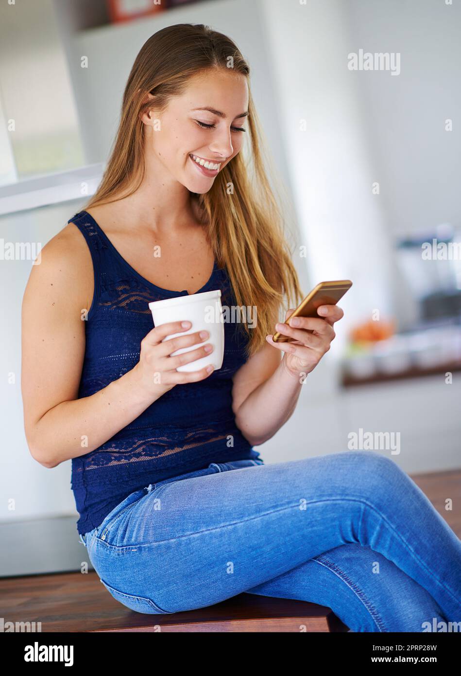 Il suo tempo per una certa azienda. Una donna seduta sul suo bancone della cucina mentre tiene il suo caffè e cellulare. Foto Stock