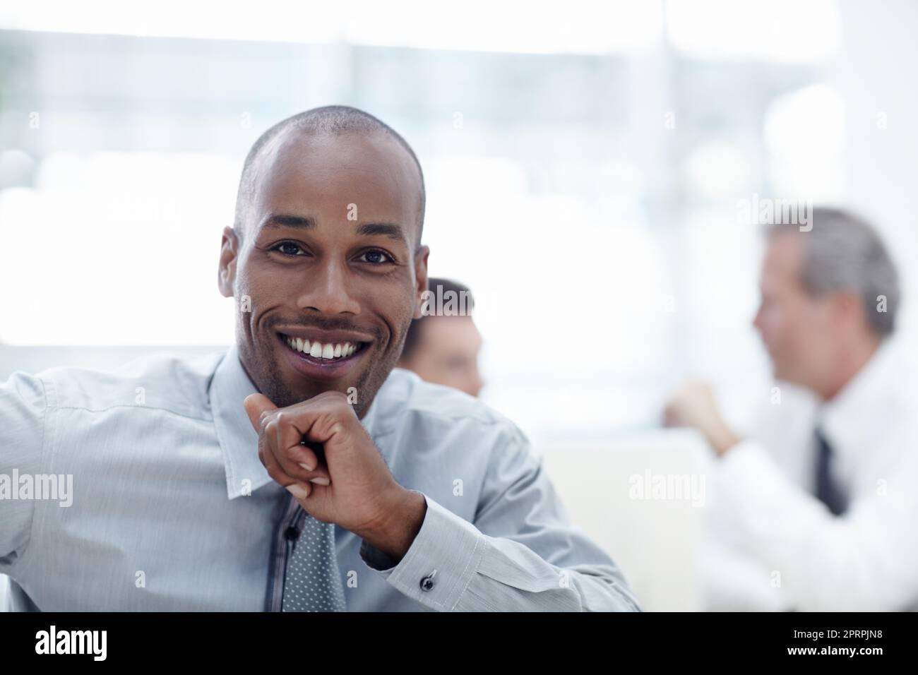 Che cosa è la vostra idea - crescere il commercio. Un bell'uomo d'affari africano che gli riposa il mento sulla mano e sorride con i colleghi sullo sfondo. Foto Stock