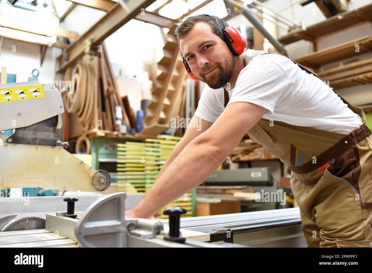 Ritratto di un falegname in abiti da lavoro e di protezione dell'udito in officina di una bottega del falegname Foto Stock