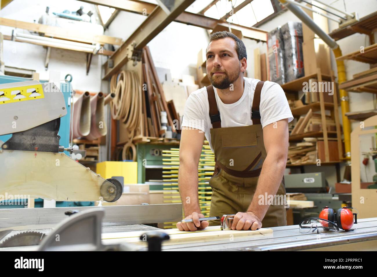 Gentile falegname con protezioni per le orecchie e gli indumenti da lavoro lavorando su una sega in officina Foto Stock