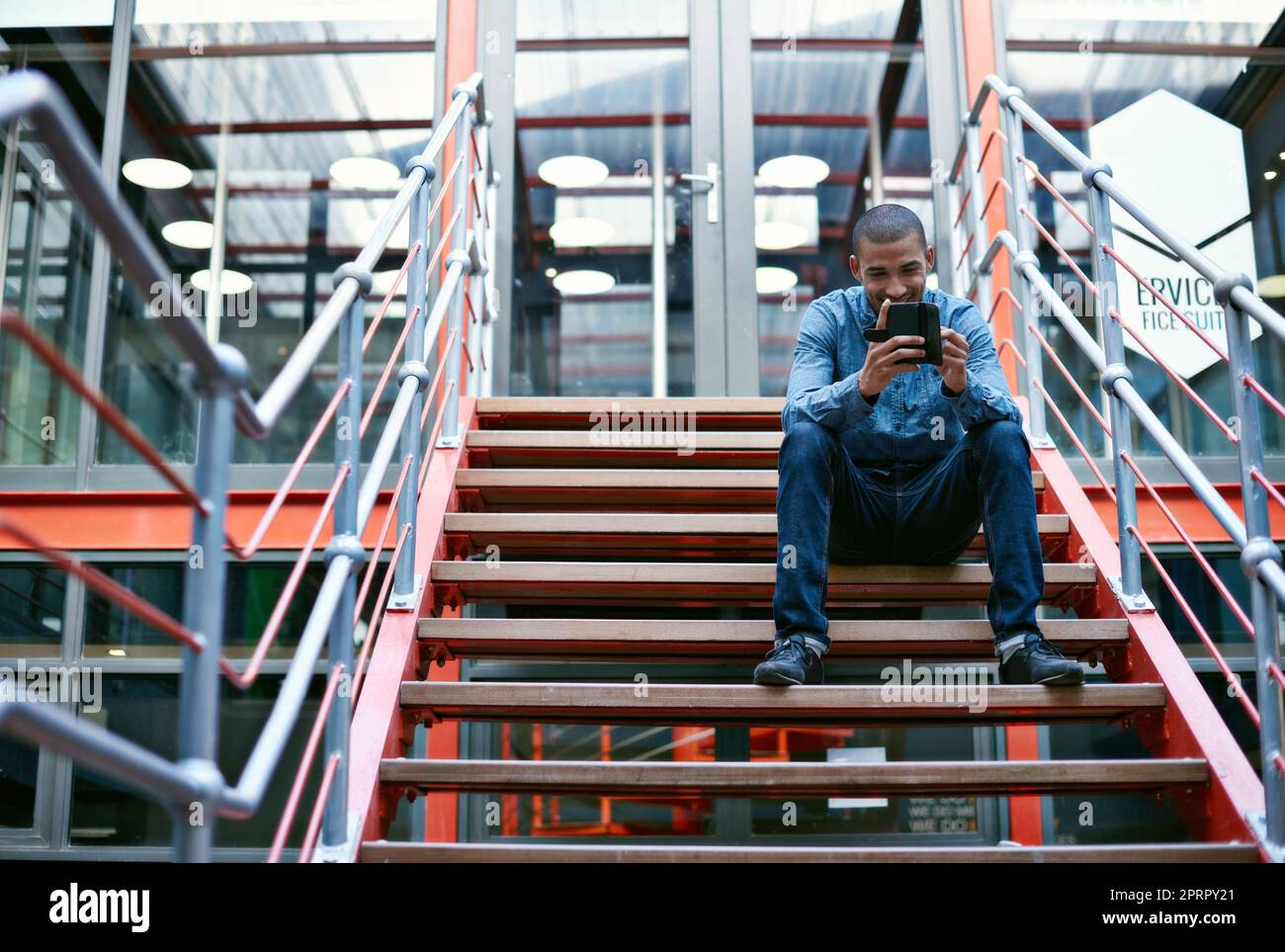 È sempre buono ottenere un feedback positivo. Un bel giovane uomo che usa il suo telefono cellulare mentre si siede sui gradini di un ufficio. Foto Stock