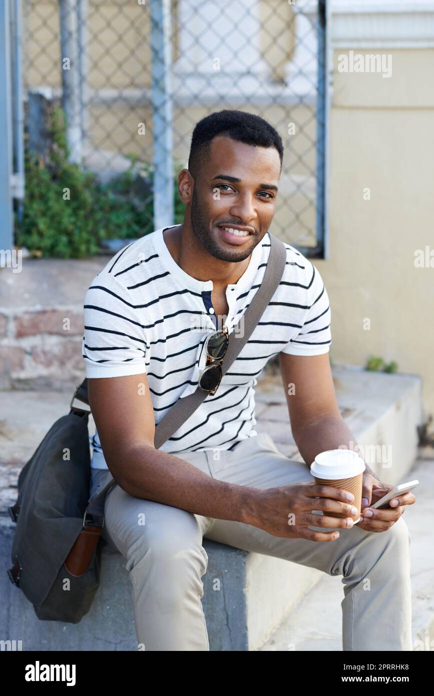 Pausa caffè in movimento. Un bell'uomo d'affari afro-americano vestito casualmente seduto in città e con il suo cellulare mentre beve il caffè. Foto Stock