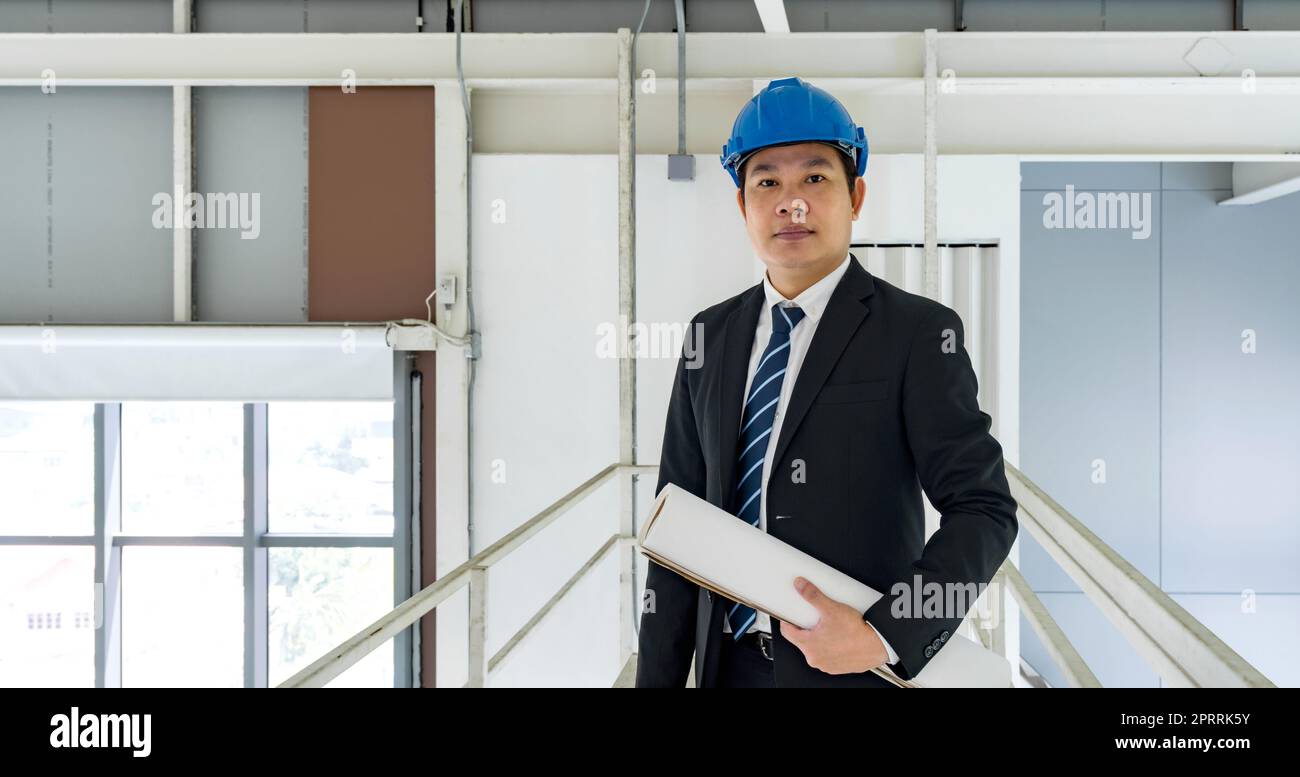 Uomo d'affari asiatico con vestito nero che indossa un cappello da costruzione blu, con disegno di costruzione. L'ispettore ispeziona l'ordine della fabbrica sulla passerella. Foto Stock