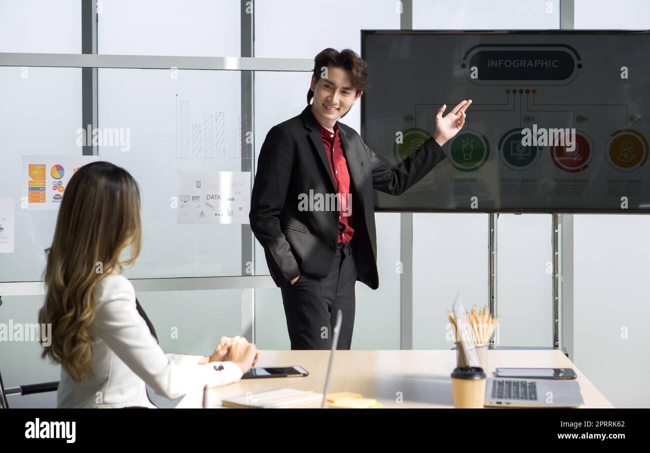 Giovane uomo d'affari asiatico in tuta che presenta con un grande monitor digitale. Riunione del team dei dirigenti aziendali in un ufficio moderno con computer portatile, tablet e caffè sul tavolo. Foto Stock