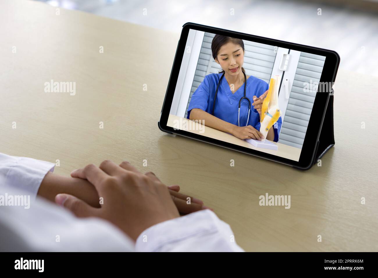 Vista posteriore di donna in abito bianco hanno conferenza webcam su tablet computer con giovane medico asiatico in uniforme blu su ossa, articolazioni, tendini e muscoli. Foto Stock