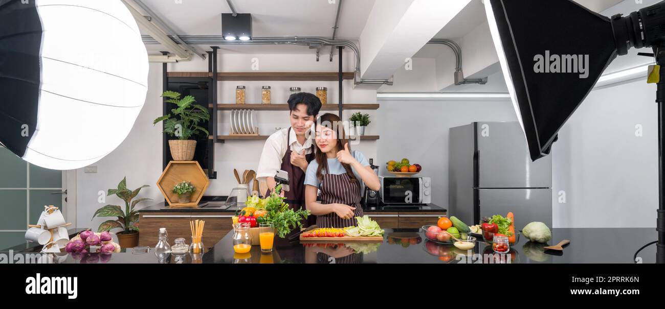 Le coppie asiatiche passano del tempo insieme in cucina. Giovane donna in grembiule che cucina insalata mentre il suo ragazzo registra video vlog per social blogger. Luci da studio fotografico allestite in cucina. Foto Stock