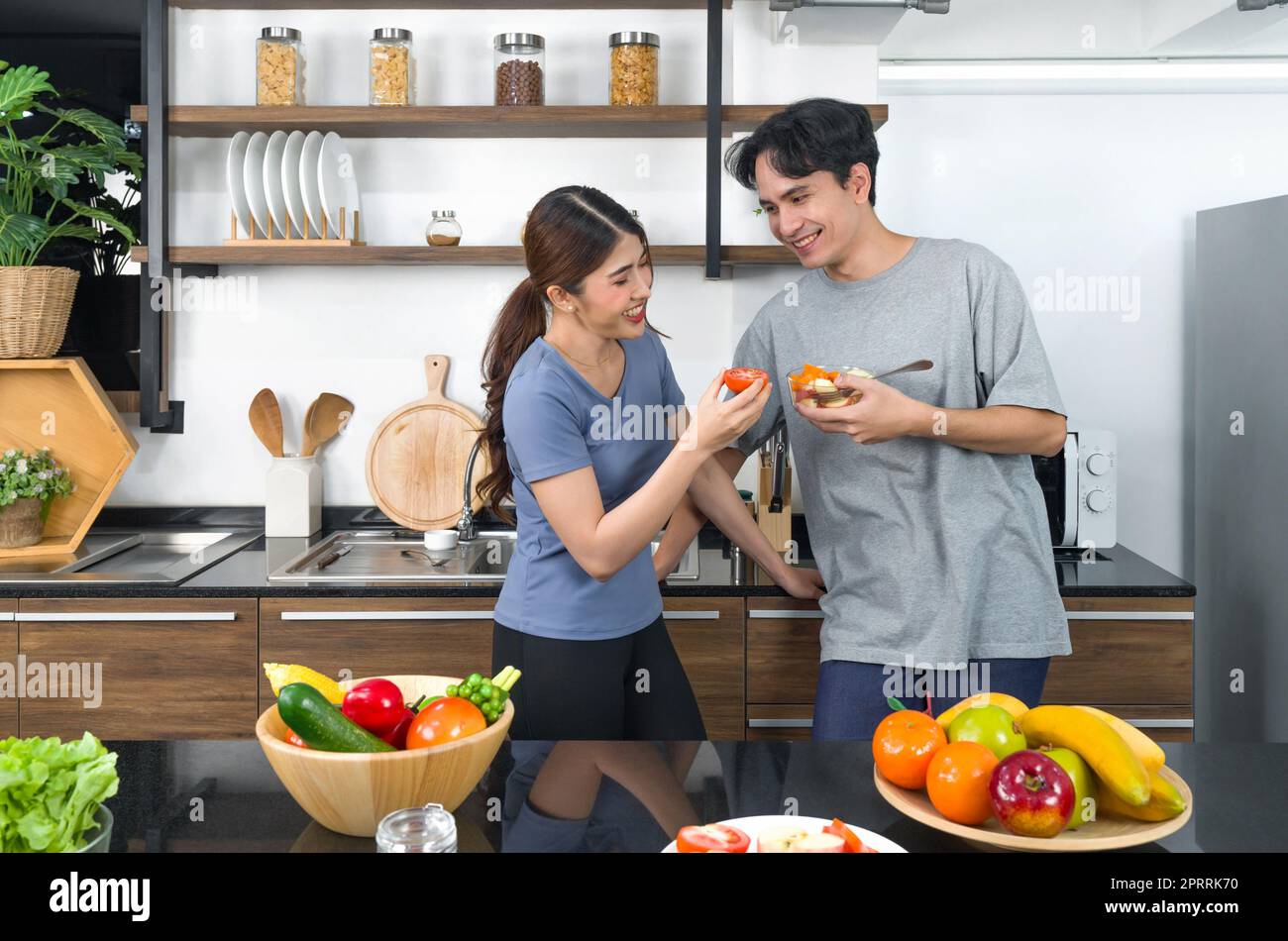 Le coppie asiatiche passano del tempo insieme in cucina. Giovane donna che mangia pomodoro mentre il suo ragazzo tiene una ciotola per insalata. Sul bancone è disponibile una varietà di frutta e verdura. Foto Stock
