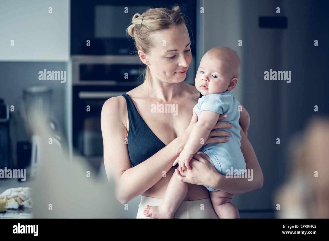 Bella giovane madre che tiene il suo bambino neonato in piedi vicino alla finestra della cucina a casa Foto Stock