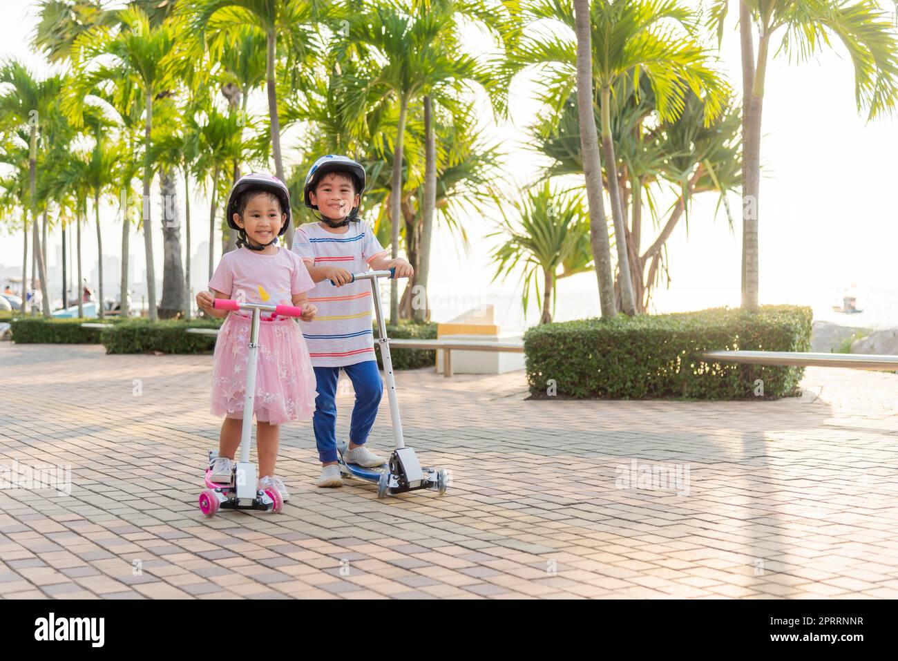I bambini e le bambine asiatiche felici indossano un casco sicuro e giocano a calcio su strada nel parco all'aperto il giorno d'estate Foto Stock
