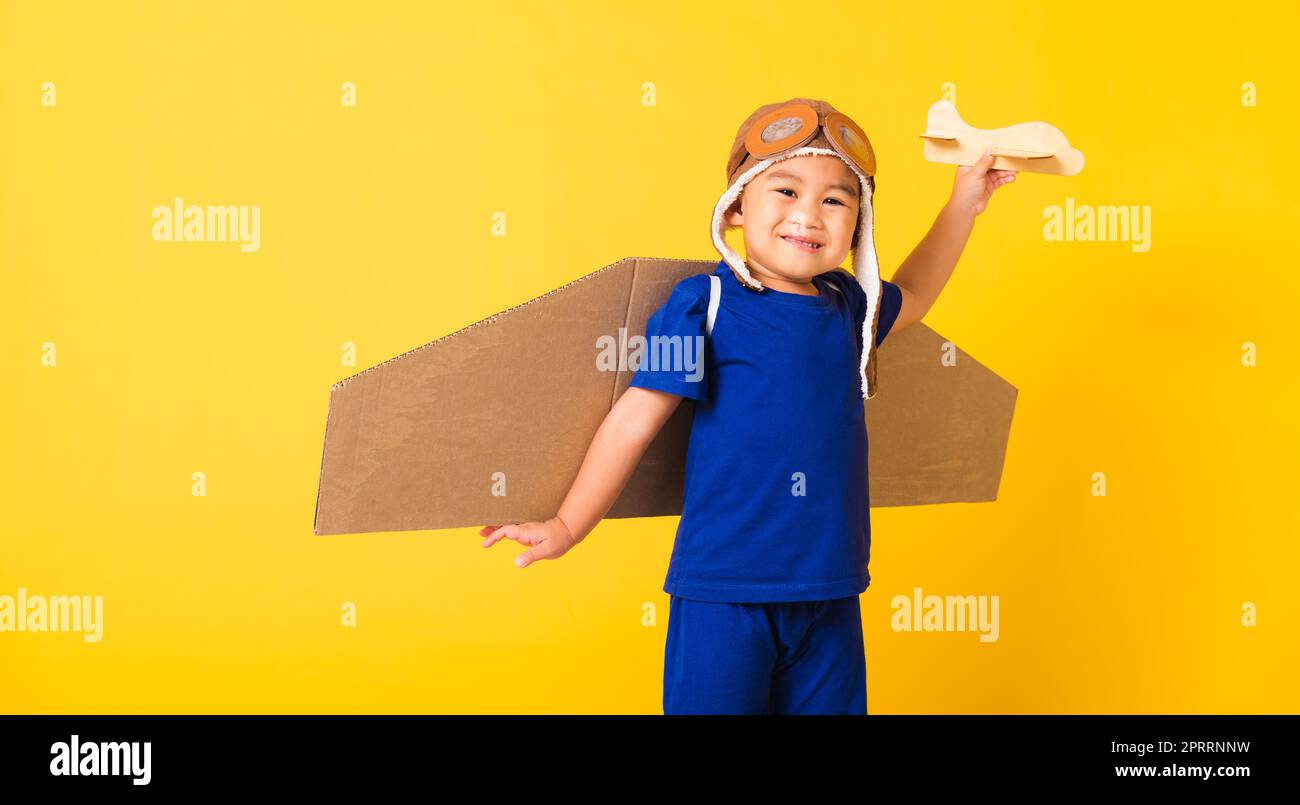 Bambino piccolo ragazzo sorriso indossare pilota cappello gioco e occhiali di protezione con ali di aeroplano giocattolo in cartone Foto Stock