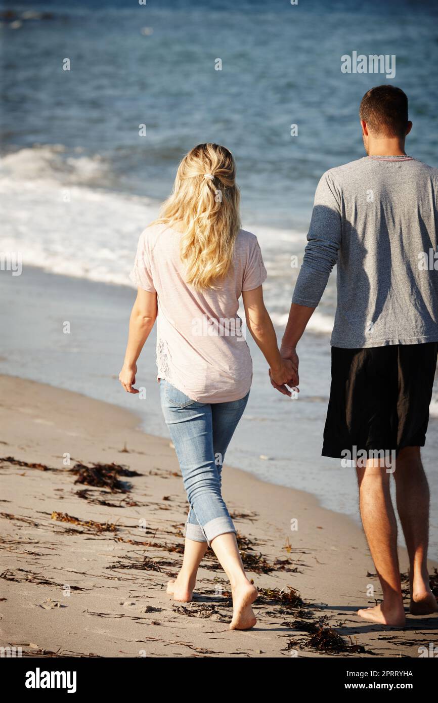 Allontanarsi da tutto. Ripresa da dietro di una giovane coppia che cammina mano in mano lungo una spiaggia sabbiosa. Foto Stock