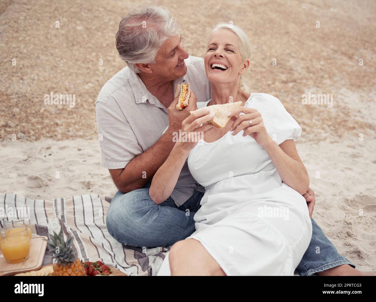 Amore e sana coppia anziana su picnic in spiaggia mangiare panino con conversazione divertente per il benessere, la salute o la pensione. Donna e uomo anziani all'aperto con pane al formaggio insieme a terra Foto Stock