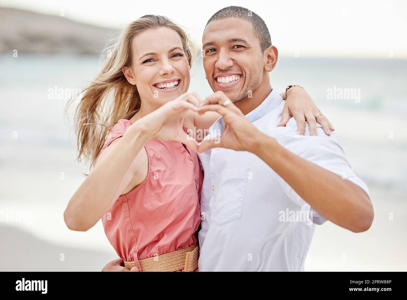 Felici coppie interrazziali mostrano il segno del cuore con le mani e si godono l'aria fresca in vacanza in spiaggia mentre si legano. Ritratto di coppia che abbraccia l'oceano con sorriso e amore insieme in vacanza Foto Stock