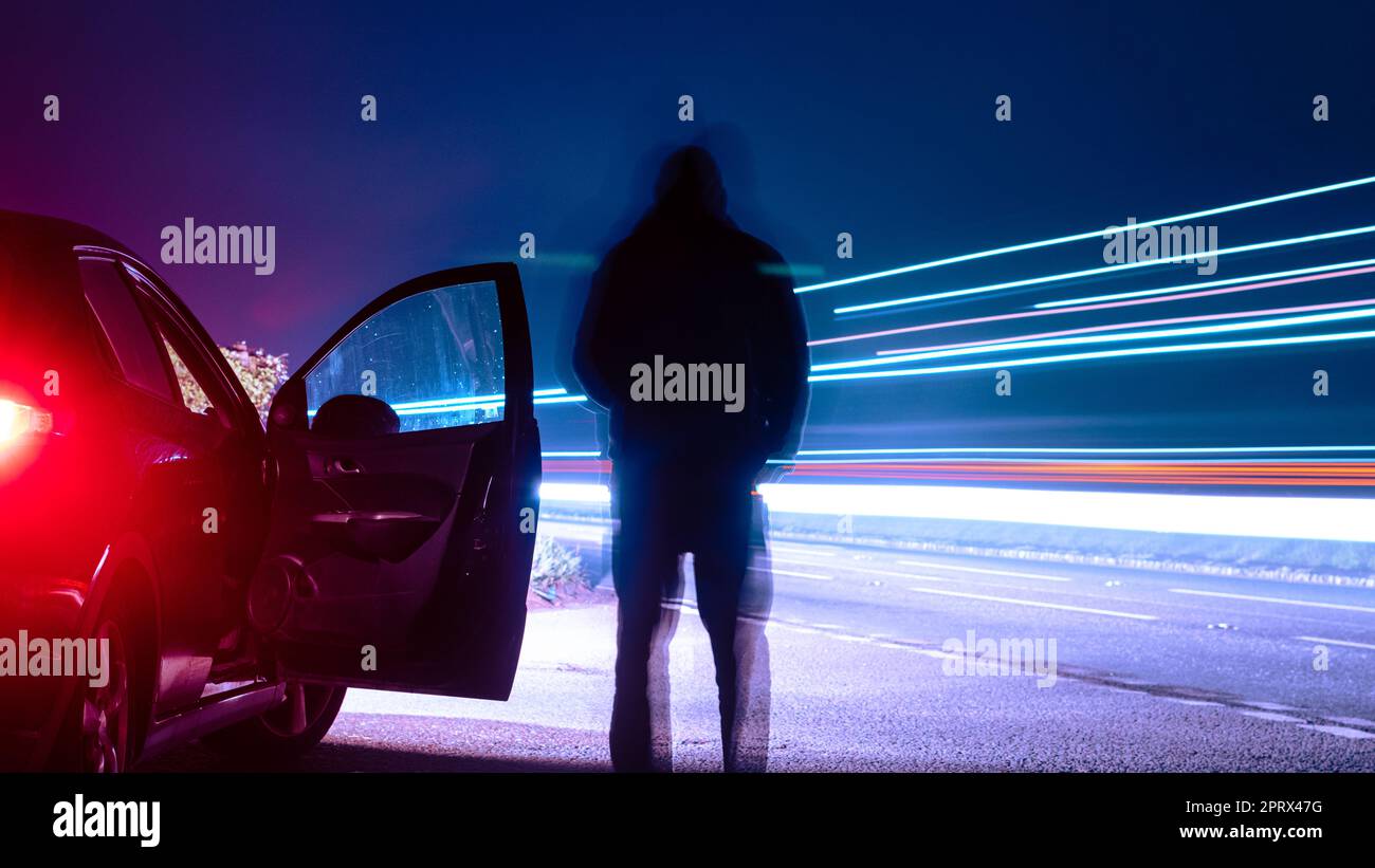 Una figura sfocata e silhouette, che guarda i sentieri del traffico. In una notte di nebbia e nebbia, accanto a un'auto. Foto Stock