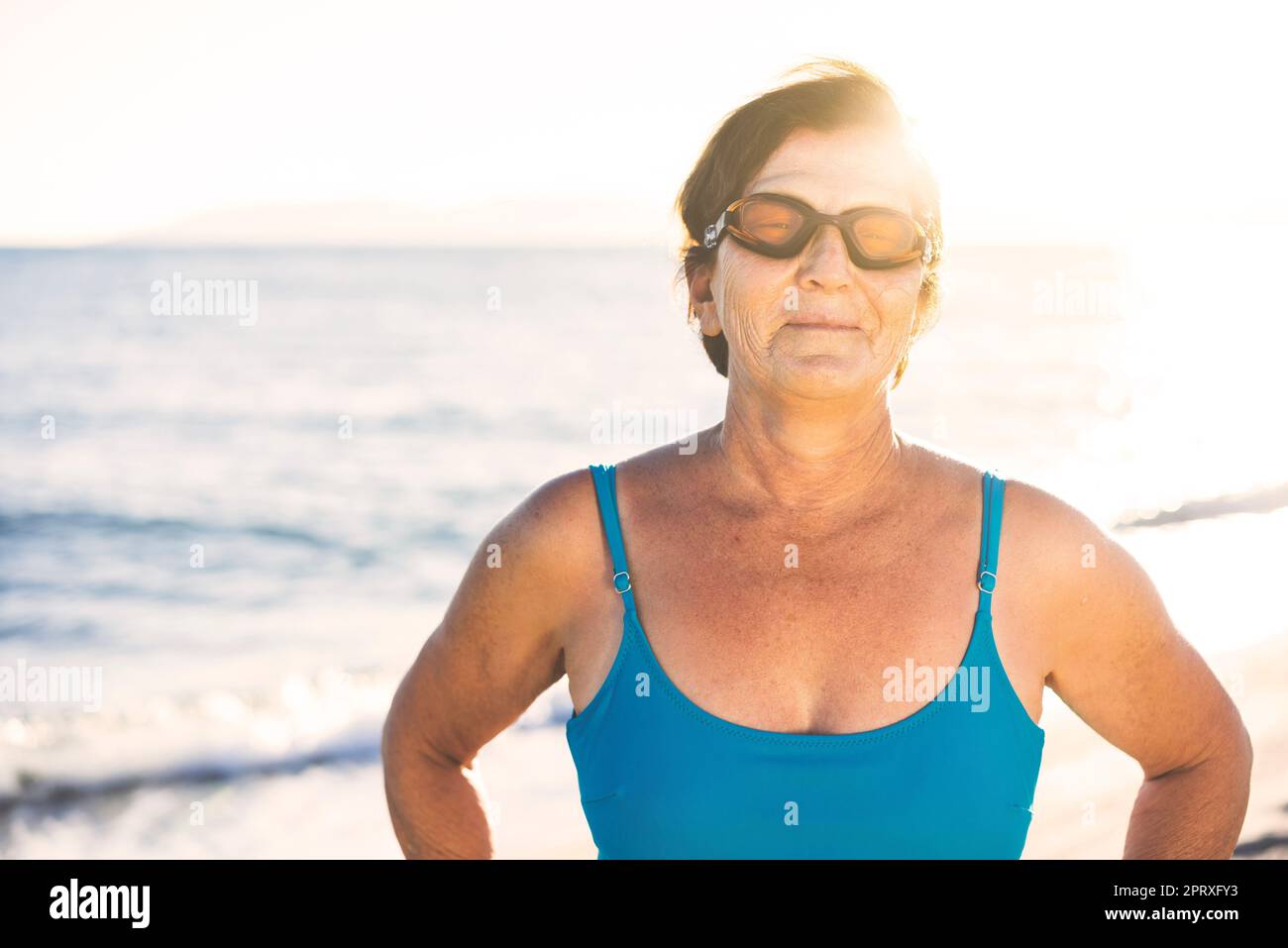 Donna anziana in spiaggia, in piedi accanto al mare. Indossa un costume da bagno blu e occhiali da nuoto, pronti per nuotare Foto Stock