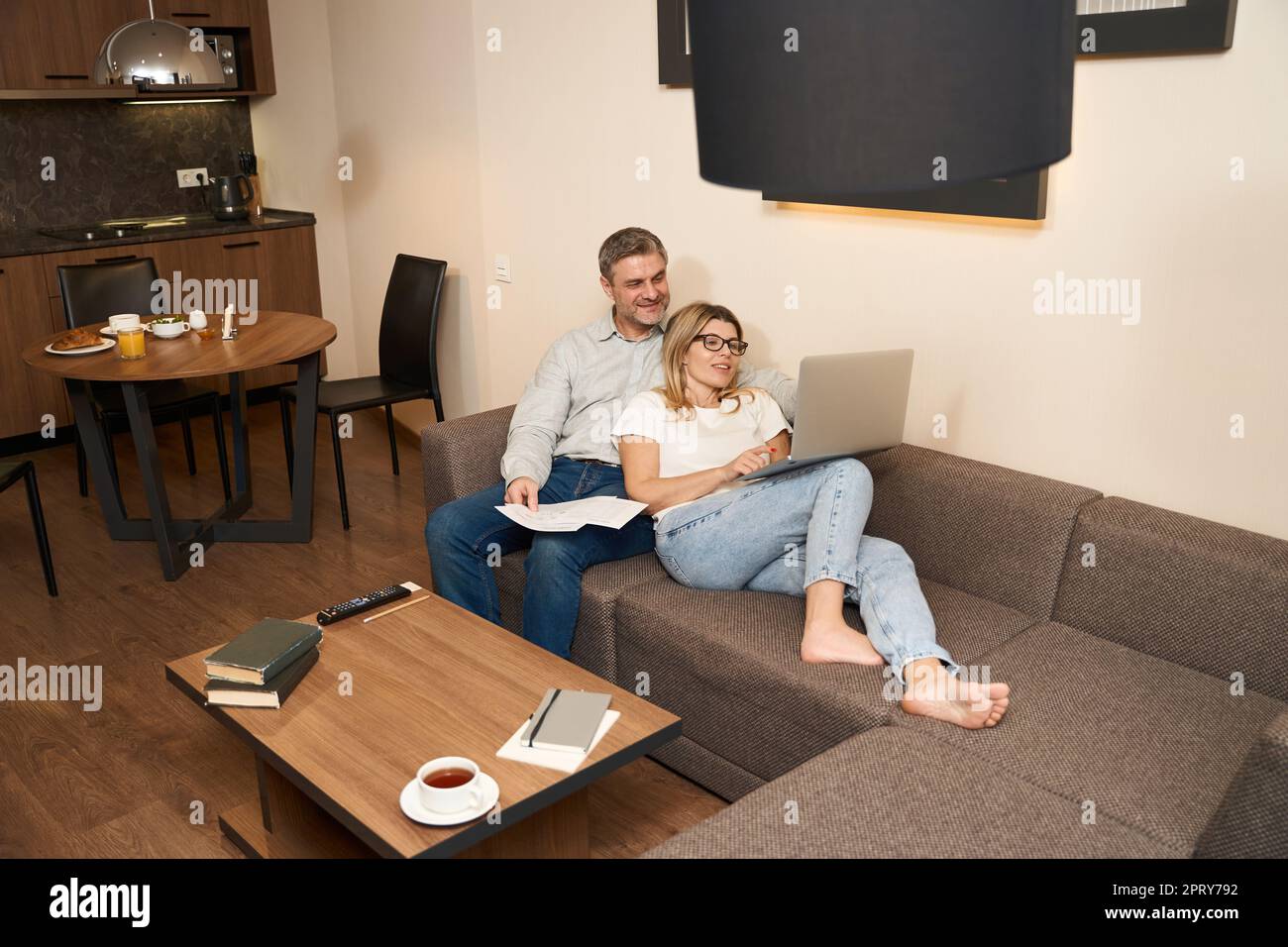 Bell'uomo seduto su un comodo divano e guardando il computer portatile di moglie che è sul suo grembo in appartamento Foto Stock