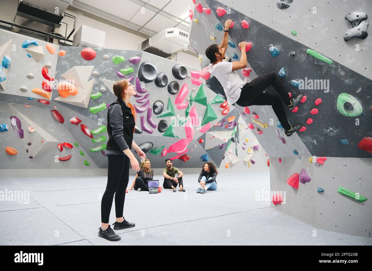 Gruppo multiculturale che guarda un amico sul muro di arrampicata nel centro di attività al coperto con pullman Foto Stock