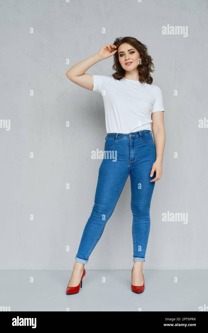 Ragazza positiva in jeans, t-shirt bianca e paletti rossi che si posano vicino alla parete tenendo la mano vicino alla testa Foto Stock