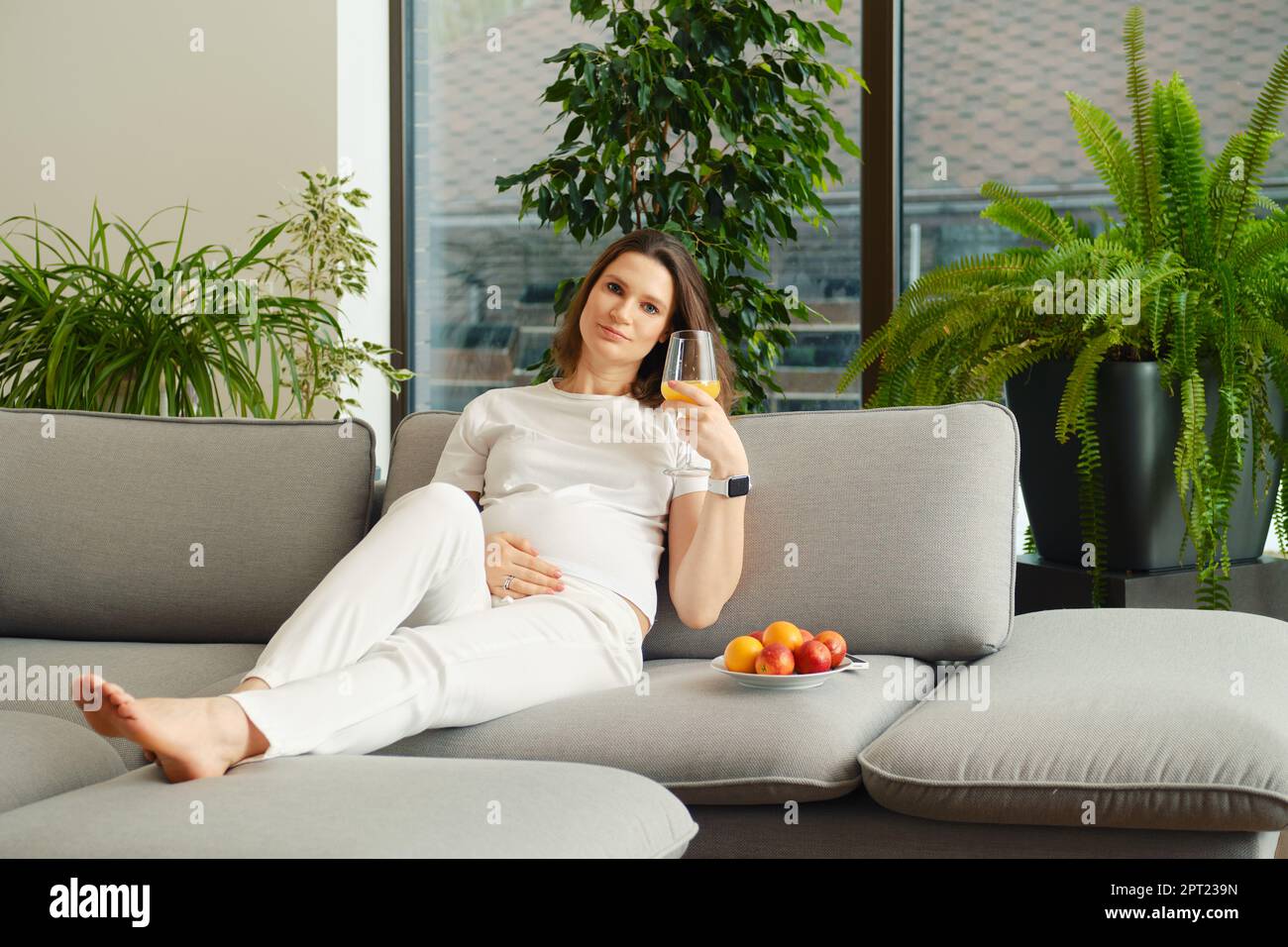 Donna incinta carina si siede sul divano con un bicchiere di succo d'arancia fresco in mano Foto Stock