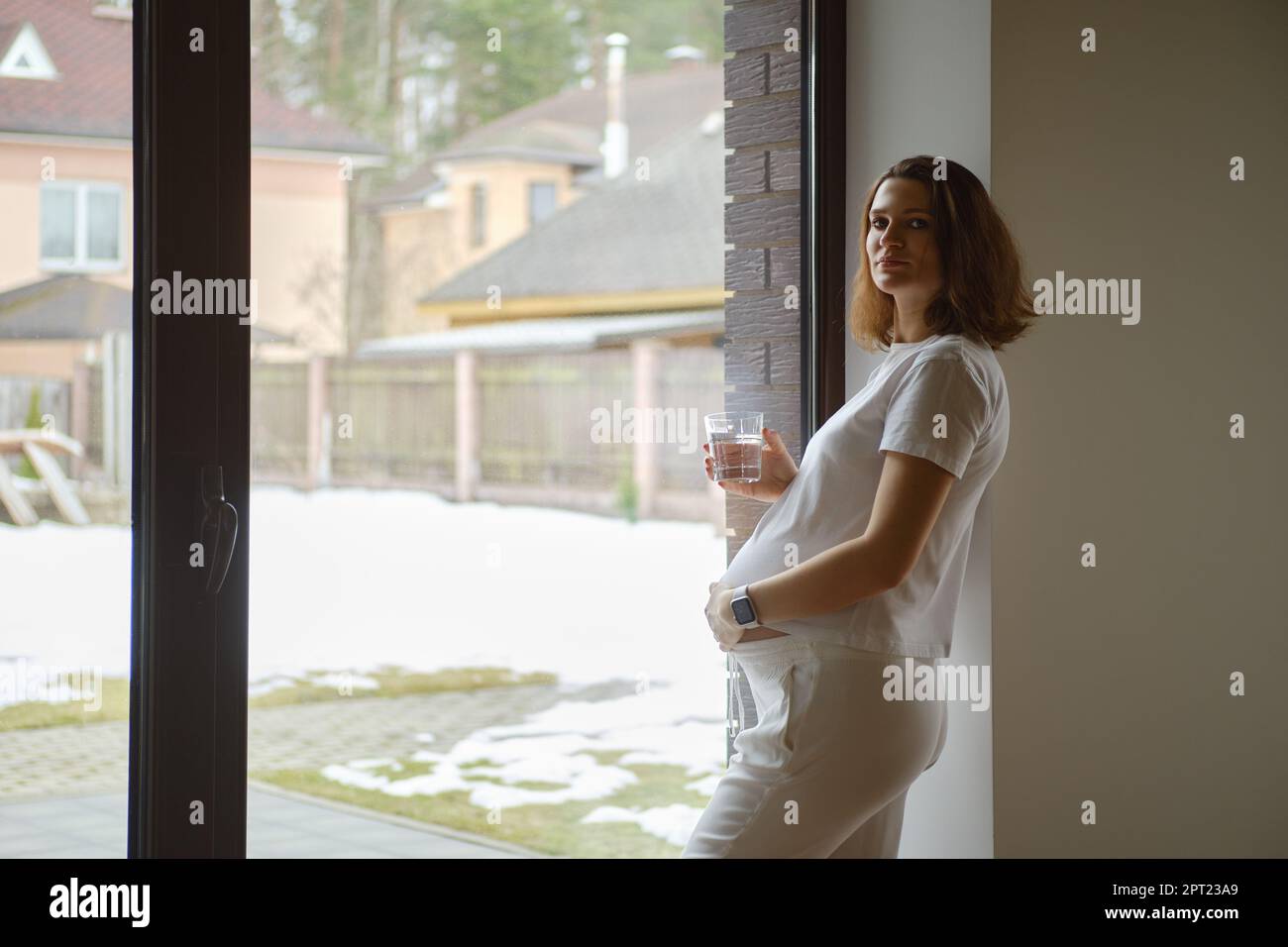 La donna incinta pensiva con un bicchiere d'acqua in mano guarda fuori dalla sua finestra della cucina appoggiata alla parete Foto Stock